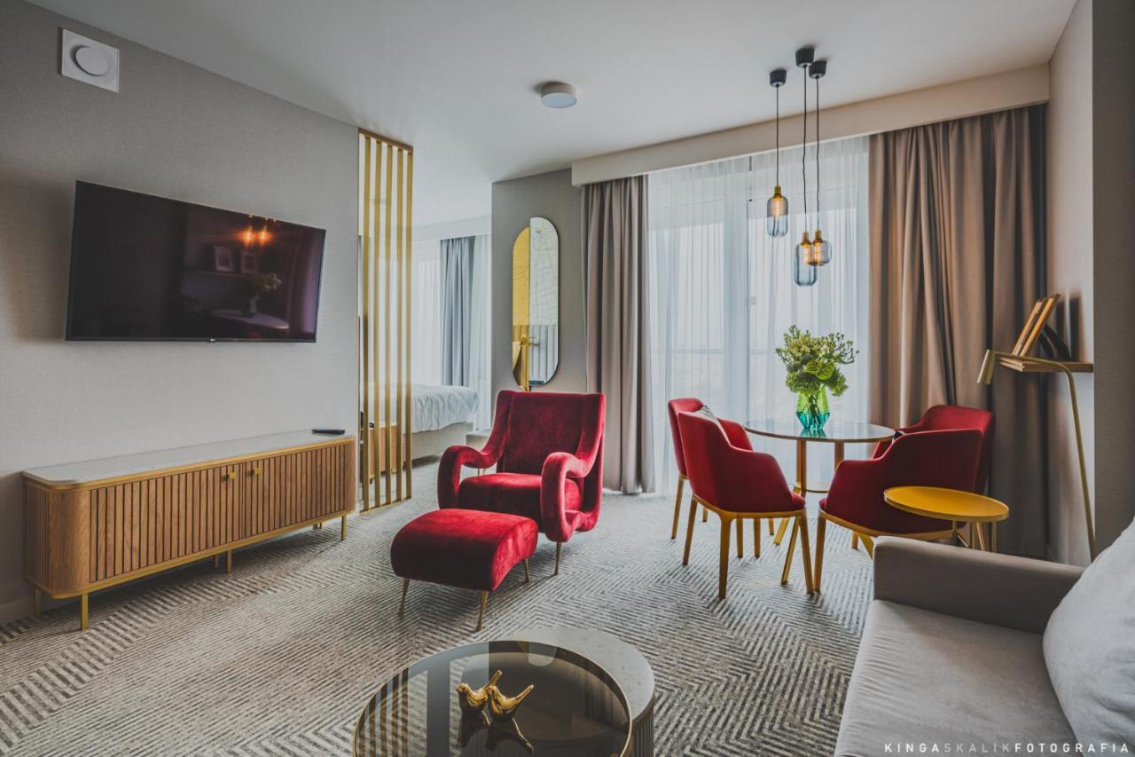 Royal Tulip Warsaw Centre Aparthotel Exterior foto The photo shows a modern and stylish living space. It features a comfortable seating area with a pair of red armchairs and a small round dining table accompanied by two red chairs. A gray sofa is also present. There is a flat-screen TV mounted on the